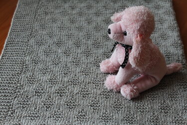 Gehäkelte graue Babydecke mit quadratischem Reliefmuster und rosa Plüschpudel