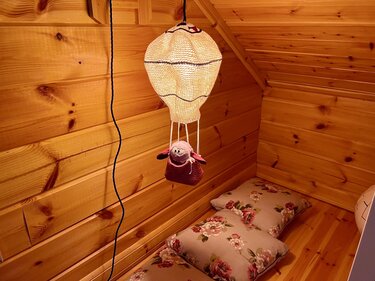 Hand-crocheted hot air balloon mobile with a small stuffed toy in a yarn basket hanging in a wooden nook above floral cushions.