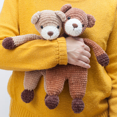 Person in mustard sweater holding two crocheted plush teddy loveys, one beige and one brown