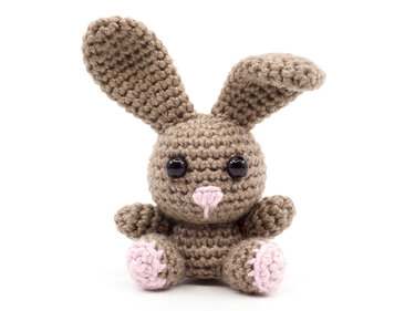 Brown crocheted amigurumi bunny with long ears, black safety eyes, and a pink embroidered nose on a white background.
