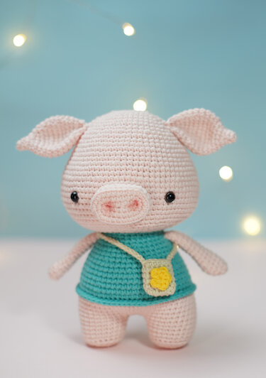 Light-pink crocheted pig amigurumi wearing a teal shirt and small yellow pouch against a soft blue bokeh background.