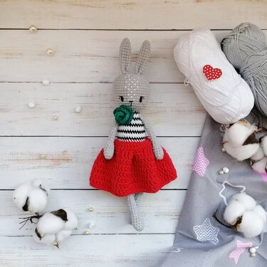 Grey crocheted amigurumi rabbit wearing a red skirt and striped top, shown with yarn and cotton.