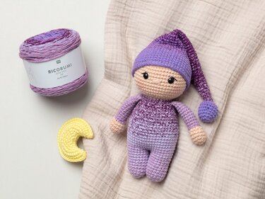 Purple-gradient amigurumi doll wearing a long pointed nightcap with pompom, next to a skein of purple yarn and a small yellow crocheted crescent moon on a beige muslin cloth.