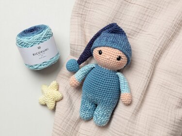 Gehäkelte Amigurumi-Schlafpuppe in Blautönen mit Zipfelmütze, daneben Knäuel Farbverlaufsgarn und gehäkelter Stern.