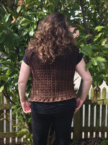 Rückenansicht einer Frau in brauner, gehäkelter ärmelloser Weste vor grüner Bepflanzung