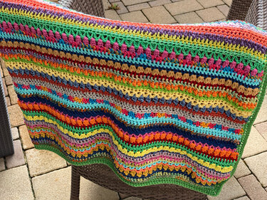 Bunte, gestreifte Häkeldecke mit unterschiedlichen Maschenstrukturen über einem Gartenstuhl.