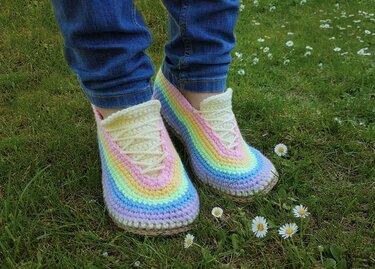 Füße in blauen Jeans tragen gehäkelte Slipper in Pastell-Regenbogenfarben mit hellen Schnürsenkeln auf grünem Gras mit Gänseblümchen.