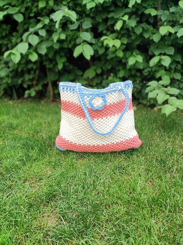 Handgehäkelter Shopper mit creme- und korallenfarbenen Streifen, hellblaue Einfassung und mittige Blüte auf Rasen