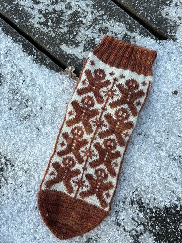 Gestrickte Socke mit braunen Lebkuchenmännchen auf hellem Grund, liegend auf verschneiter Holzfläche