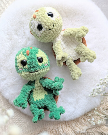 Zwei flauschige gehäkelte Amigurumi‑Schildkröten in Grün auf weißem Plüschuntergrund