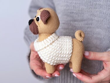 Beige crocheted pug amigurumi wearing a cream sweater held in hands.