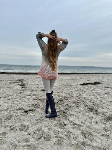 Rückenansicht einer Frau am Strand in langem gehäkelten Pullover mit Ombre-Farbverlauf und offenem Lochmuster