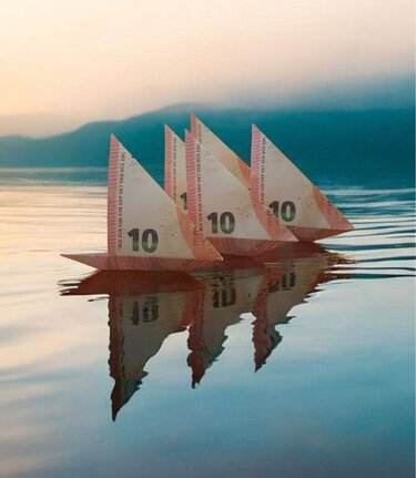 Reihe von vier Origami-Booten aus 10-Euro-Scheinen auf ruhigem Wasser mit klarer Spiegelung.