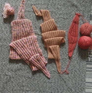 Three ribbed knitted triangle shawl pieces in pink, beige and coral with yarn balls on textured fabric.