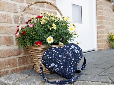 Herzförmige dunkelblaue Stofftasche mit floralem Druck und Schulterriemen neben einem Weidenkorb voller Blumen.