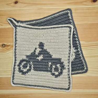 Zwei gehäkelte quadratische Topflappen in beige und grau, einer mit dunkelgrauer Motorrad-Silhouette und kontrastierendem Rand, auf Holzuntergrund
