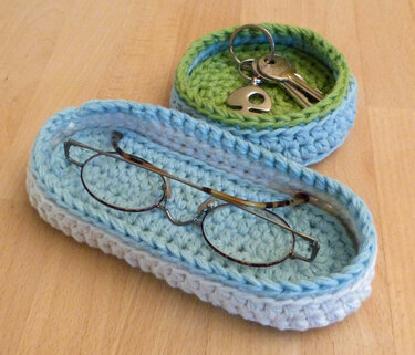 Gehäkeltes ovales Körbchen mit Brille und rundes Körbchen mit Schlüsseln in Blau, Grün und Grau auf Holztisch