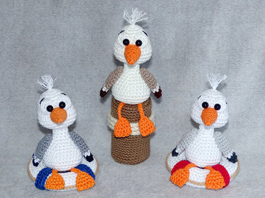 Drei gehäkelte Amigurumi‑Möwen vor grauem Hintergrund; eine große Möwe auf einem Zylinder, zwei kleine auf Rettungsringen.