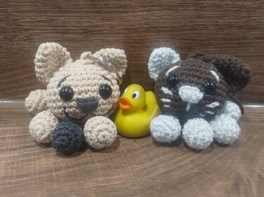 Beiges Amigurumi mit Schlüsselring, braunes Amigurumi mit weißen Akzenten und eine gelbe Quietscheente auf Holz