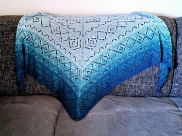 Dreieckstuch gestrickt mit Ajour‑Rautenmuster, Farbverlauf von hell- zu dunkelblau, auf grauem Sofa drapiert