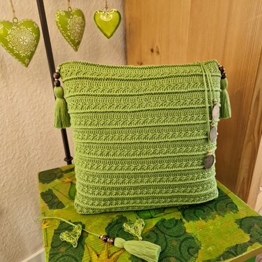 Quadratisches grünes gehäkeltes Kissen mit Quasten, Holzperlen und runden Anhängern auf grün gemustertem Tisch