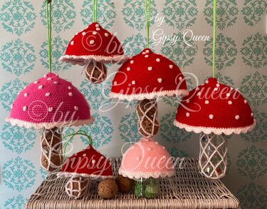 Gehäkelte Pilze in Rot und Rosa mit weißen Punkten, teils hängend an grünen Schnüren, teils stehend auf einem geflochtenen Korb vor gemusterter Tapete.