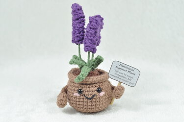 Gehäkelter brauner Blumentopf mit zwei violetten Lavendelblüten, grünen Blättern, lachendem Gesicht und kleinem Schild