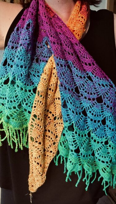 Close-up of a person wearing a multicolored crocheted triangle shawl with openwork lace pattern and fringed edge.