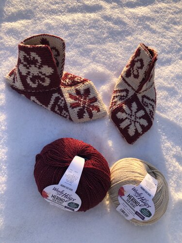 Modulare gestrickte Stulpen aus Quadraten neben zwei Garnknäueln in Rot und Beige auf Schnee