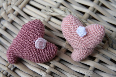 Zwei gehäkelte Herzen in Rosa und Altrosa, verziert mit kleinen Stoffrosen, auf geflochtenem Untergrund.