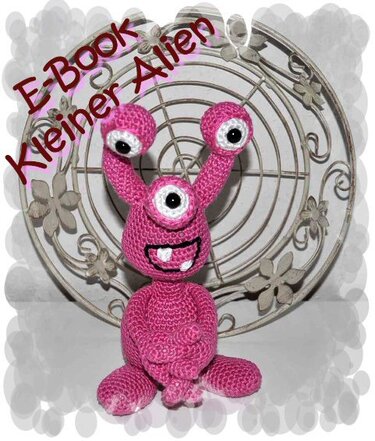 Rosa gehäkeltes Amigurumi mit drei Augen vor dekorativem Hintergrund; eingeblendeter Text 'E-Book Kleiner Alien'.