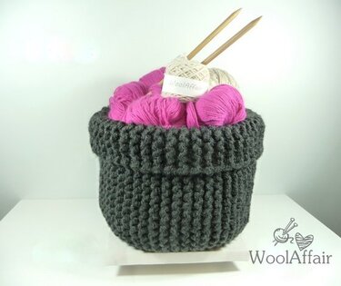 Gestrickter dunkelgrauer Korb mit pinker und cremefarbener Wolle und zwei Holzstricknadeln auf hellem Hintergrund.