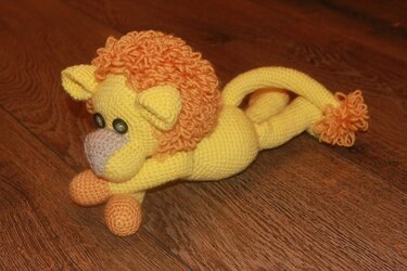 Gehäkelter gelber Amigurumi-Löwe mit orangefarbener Mähne und Knopfaugen auf Holzboden