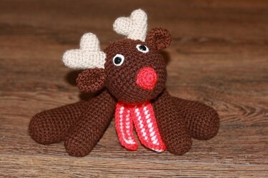 Gehäkeltes braunes Rentier-Amigurumi mit beigen Geweihen, roter Nase und rot-weißem Schal auf Holzboden