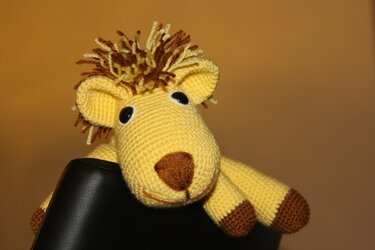 Gelb-brauner gehäkelter Amigurumi-Löwe mit fransiger Mähne, liegt über einer schwarzen Stuhllehne
