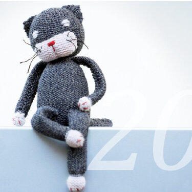 Gestrickte graue Amigurumi-Katze mit weißer Schnauze und roter Nase, sitzend auf einer hellen Fläche.