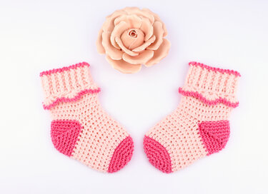 Paar gehäkelter Babysocken hellrosa mit dunkelpinkem Zehen- und Fersenbereich neben rosafarbener Blüte