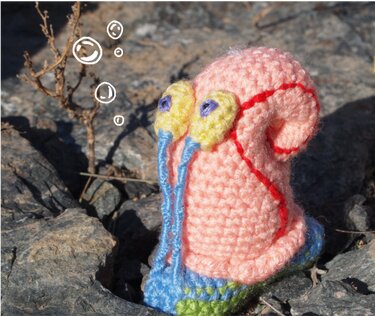 Gehäkelte Amigurumi‑Schnecke mit rosa Gehäuse, gelben Augen und blauen Fühlern auf Felsen
