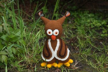 Braune handgehäkelte Amigurumi-Eule mit weißen Augenringen, orangefarbenem Schnabel und gelben Füßen auf Gras