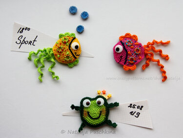 Zwei gehäkelte Fische (gelb-grün und pink-orange) und ein grüner gehäkelter Frosch mit kleiner Sternkrone auf weißem Hintergrund, dazu drei kleine blaue Häkelkreise.