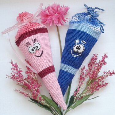 Zwei gehäkelte Schultüten, eine rosa und eine blau, mit aufgenähten Augen, Spitzenabschluss und Schleifenband, auf weißem Hintergrund mit rosa Blumen