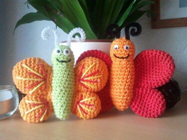 Zwei gehäkelte Amigurumi‑Schmetterlinge aus Garn in Grün, Orange und Rot neben einem Blumentopf