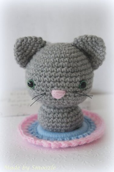 Gehäkelter grauer Amigurumi-Mäusekopf mit rosa Nase und grünen Augen auf runder Unterlage
