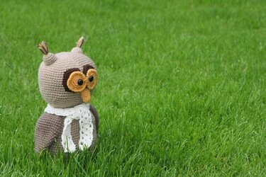 Gehäkelte Amigurumi-Eule in Braun mit weißem Schal auf grünem Rasen.