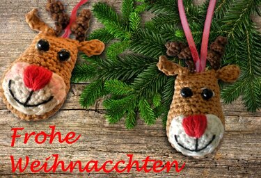 Zwei handgehäkelte Rentierköpfe mit roten Nasen, schwarzen Knopfaugen und braunen Geweihen auf Holz mit Tannenzweigen und rosa Bändern.