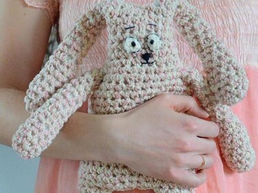 Hand hält einen gehäkelten Hasen in Rosa und Creme mit großen Augen und langen Ohren.