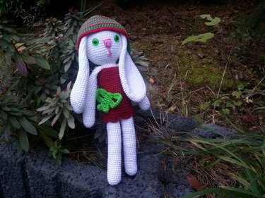 Amigurumi-Hase mit weißem Körper, rotem Kleid, grüner Schleife und gestreifter Mütze auf dunklen Steinen