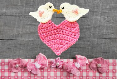 Zwei weiße gehäkelte Vögel mit gelben Schnäbeln berühren sich über einem rosa Häkelherz; darunter rosa Stoffblumen und Schleifen auf kariertem Stoff.
