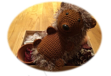 Gehäkelter brauner Igel mit zotteligem Fell in einer Holzkiste mit Zapfen und Potpourri.