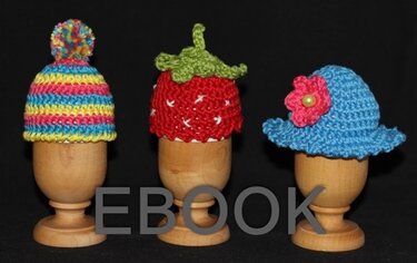 Drei gehäkelte Eierwärmer auf Holzeierbechern: bunte Bommelmütze, rote Erdbeere und blauer Sonnenhut mit Blüte